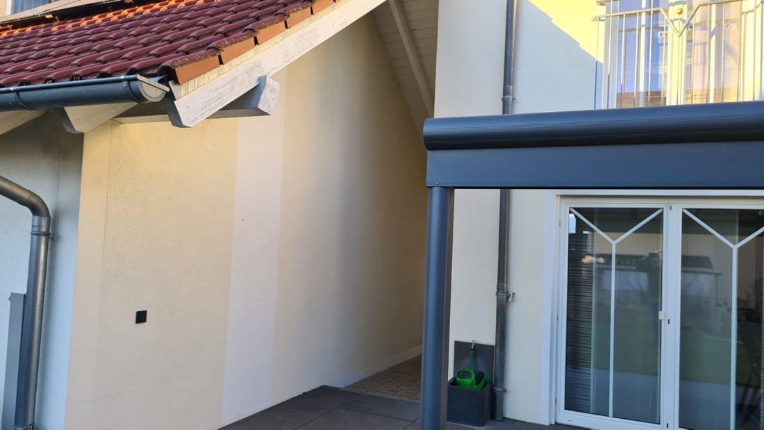 Detail einer hellen Fassade mit anthrazitfarbener Dachrinne und Terrassenüberdachung