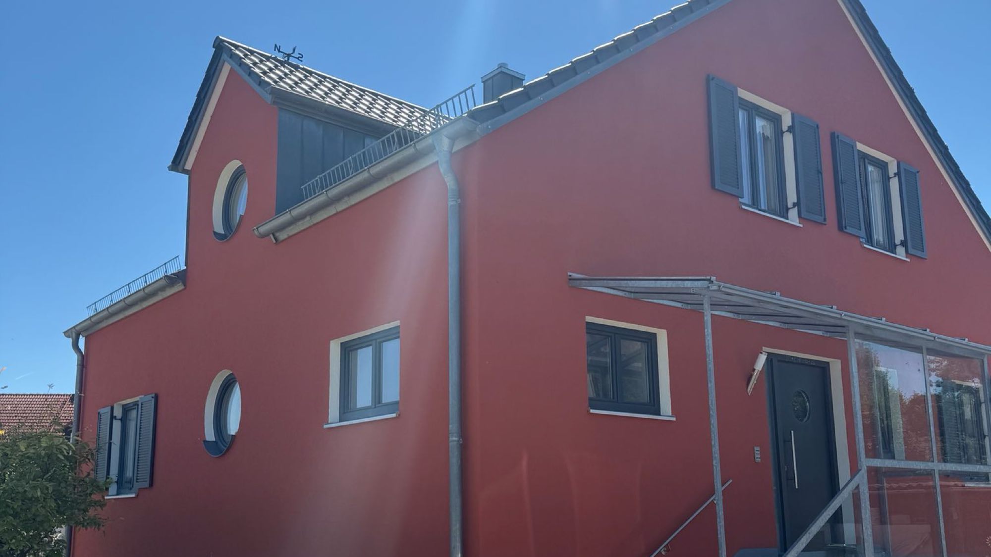 Wohnhaus mit frisch gestrichener Fassade in kräftigem Rot, anthrazitfarbenen Fensterläden und hellem Sockel vor blauem Himmel